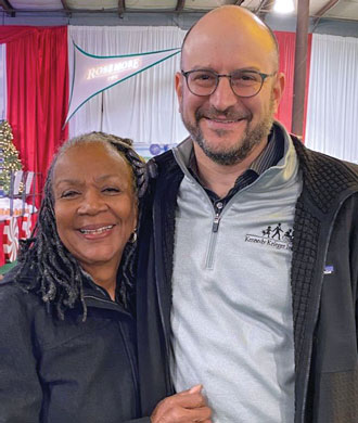 Two smiling people—a man and a woman, with their arms around each other’s backs—smile for the camera.