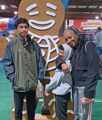Two boys, one likely a teenager and the other a younger boy, pose with a woman who has her arm around the shoulders of the younger boy. The woman and older boy are smiling. The younger boy wears a face mask. Behind them is a giant gingerbread person cut-out.