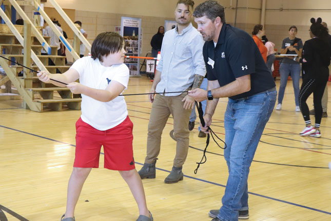 A boy holds onto the middle of a taut black rope that is connected to something out of the frame of the photo, while a man in a black polo shirt with the Whiting-Turner logo on it holds the end of the rope and gives instructions to the boy. Other children and adults stand behind the boy and man, doing similar activities.
