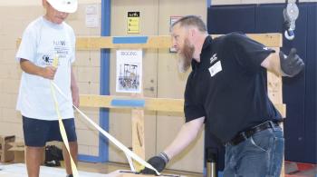 A boy wearing a construction helmet holds a rope attached to a wooden frame. A man in a black polo shirt with the Whiting-Turner logo on it holds the wooden frame and gives instructions to the boy.