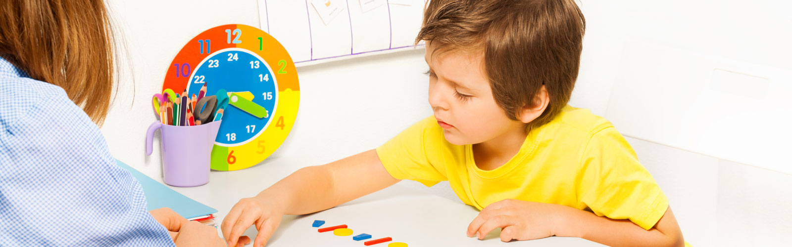 Woman teaches young child. There is a small, colorful clock in the background.