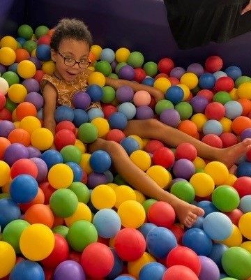 A young girl sits in a ball pit and smiles.