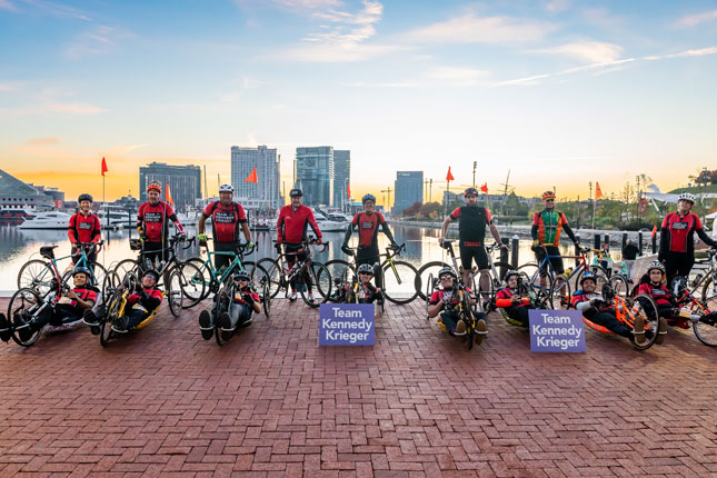 Bicyclists and hand cyclists wearing Team Kennedy Krieger apparel line up behind two purple signs that read Team Kennedy Krieger. Baltimore's skyline and inner harbor are in the background.