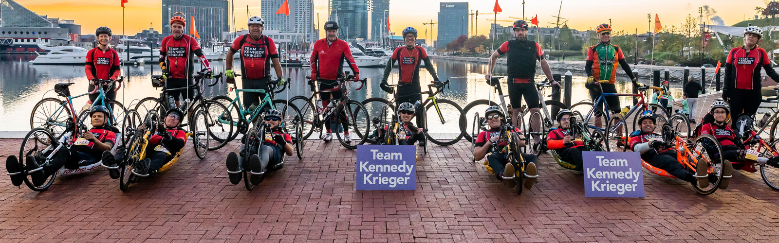 Bicyclists and hand cyclists wearing Team Kennedy Krieger apparel line up behind two purple signs that read Team Kennedy Krieger. Baltimore's skyline and inner harbor are in the background.