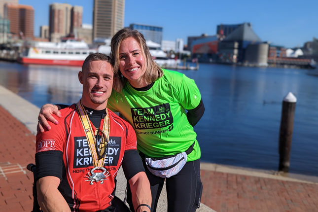 A man and a woman wearing Team Kennedy Krieger shirts at the Baltimore Running Festival. The woman is standing with her arm on the man's shoulder as he sits in a mobility device. Both are smiling.