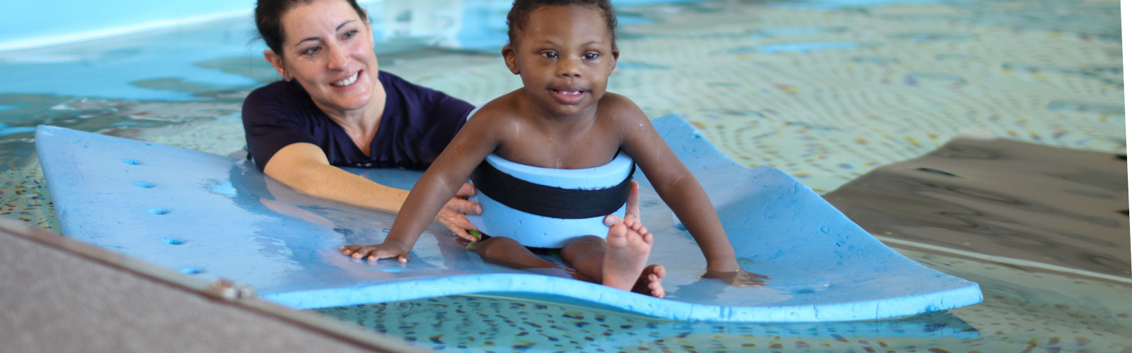 A female therapist holds a young boy around the waist as he sits on a raft during an aquatic therapy session.