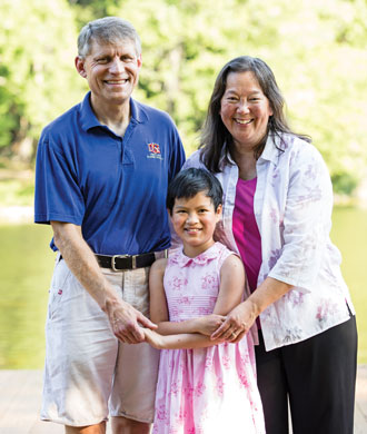 A father, mother and their young daughter smile while standing outdoors for a photo.