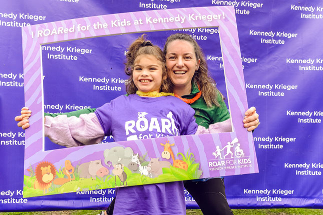 A woman and a young girl smile in front of a purple and white Kennedy Krieger Institute step and repeat banner. They are holding a frame that reads I ROARed for the Kids at Kennedy Krieger!