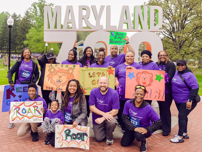 The BGE team stands in front of the Maryland Zoo sign at ROAR. They are wearing purple shirts, with some members holding hand-drawn signs with words of encouragement for runners.