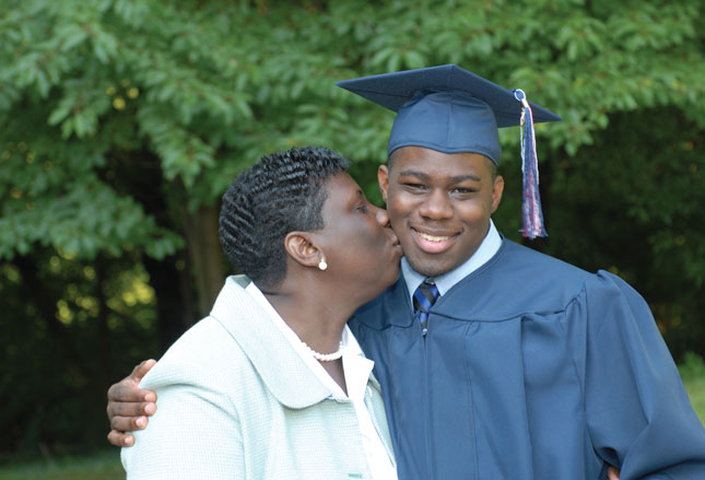 A young man wearing a cap and gown smiles while being kissed on the cheek by his mother.