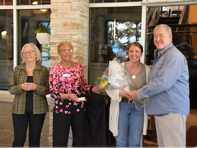 Three women and one man pose for a photo. The man is standing to the far right, presenting the woman next to him with a bouquet of flowers.