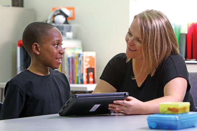 A teacher smiles while working with her student as they sit at a table. She is holding a tablet and pointing to the screen as she speaks with him.