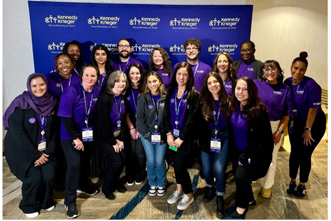 The Neurodiversity at Work team poses for a photo in front of a purple and white step and repeat banner. The banner features the Kennedy Krieger Neurodiversity at Work logo.