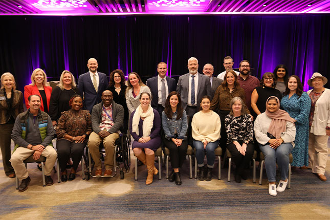 A group photo of attendees of the 2024 Neurodiversity in the Workplace Conference. Some individuals are standing in the back, behind a row of eight people who are seated.