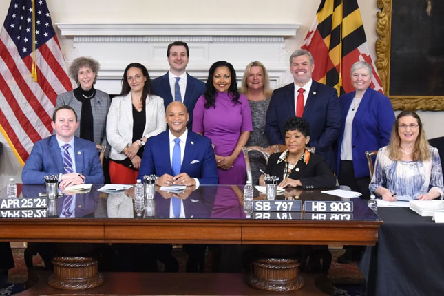 A group of people stand behind governor Wes Moore at the signing ceremony for Education - Access to Attorneys, Advocates, and Consultants for Special Education Program Fund." A man is sitting to Governor Moore's right, while two women sit to his left.