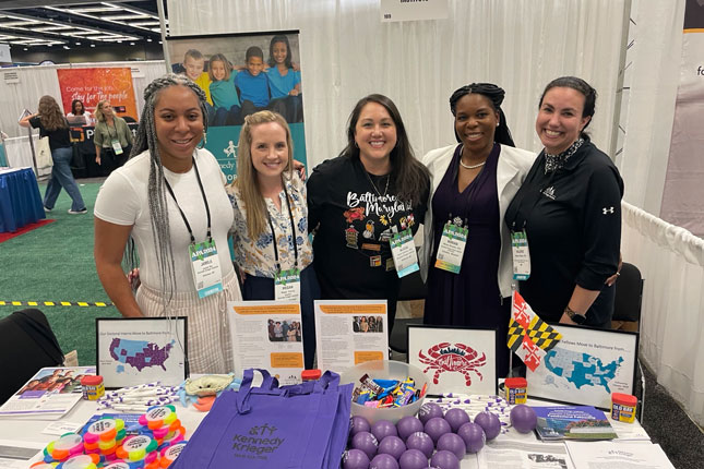 Five women stand and smile for a photo at MCDD's exhibitor table at the APA Conference.