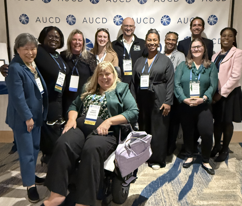 A large group of people pose in front of an AUCD step and repeat banner. One woman in the photo is sitting her mobility device, the others are standing.