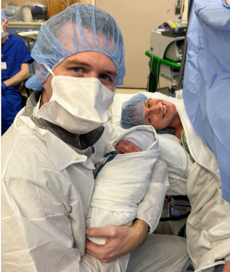 Mallory, Matt and Cora Finn Legg in the delivery room. Matt is holding Cora as Mallory lays behind them and smiles.