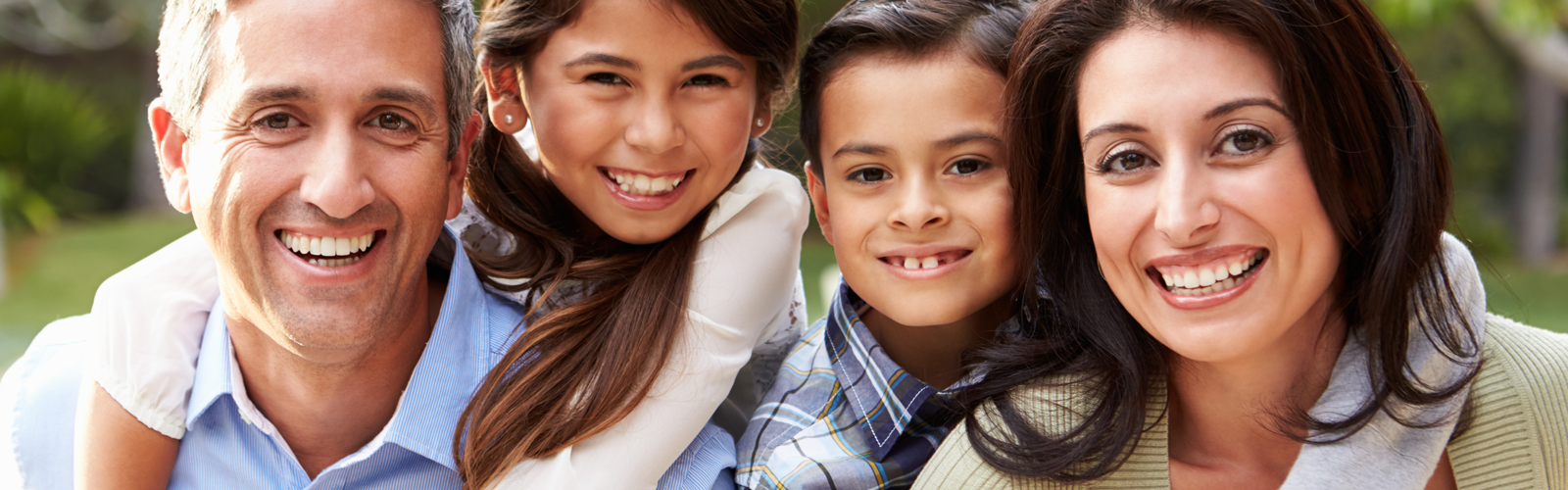 A father, mother, daughter and son smile in a family portrait that is taken outside on a sunny day.