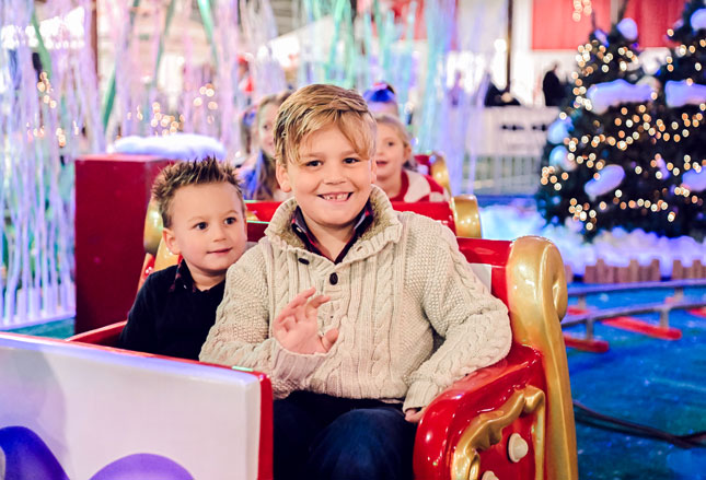A boy smiles and waves while riding a ride at Festival of Trees. A younger boy is seated on the ride alongside him.