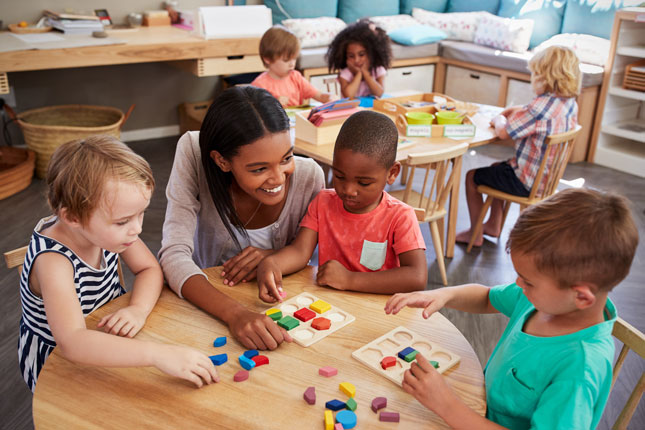 A teacher smiles as she helps three pre-school students with an activity at their desk.