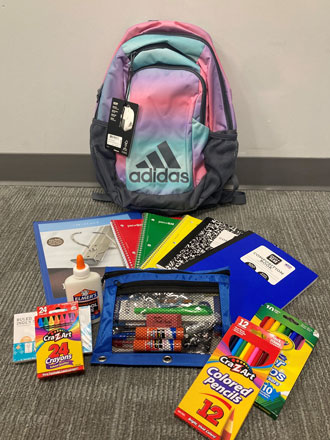 A multicolored backpack sitting on the floor, surrounding by school supplies.