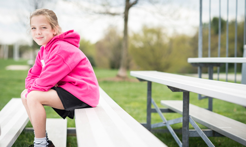 A young girl wearing a pink sweatshirt and a pair of black gym shirts sits on aluminum bleachers.