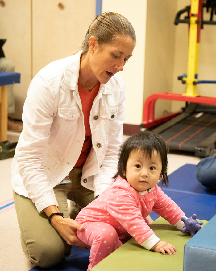 A young patient looks at the camera while a physical therapist holds her leg during a therapy session.