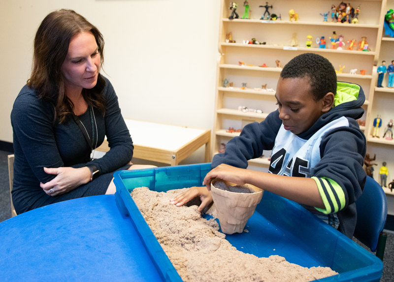 A boy plays with sand while a social worker looks on.