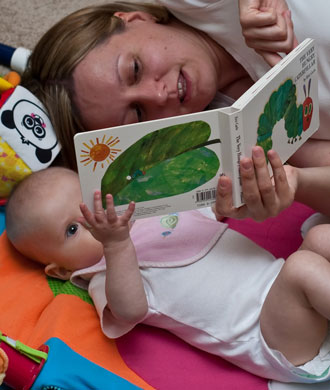 A mother lays on the ground next to her baby, holding and pointing to pages inside of the Hungry Little Caterpillar.