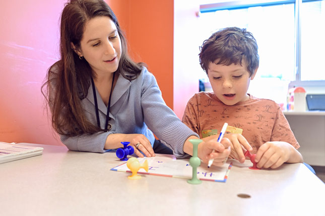 An SLP smiles as she does speech therapy with a young boy.