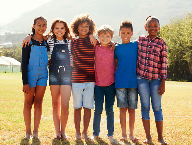 Six pre-teen friends stand shoulder to shoulder while smiling in a park.