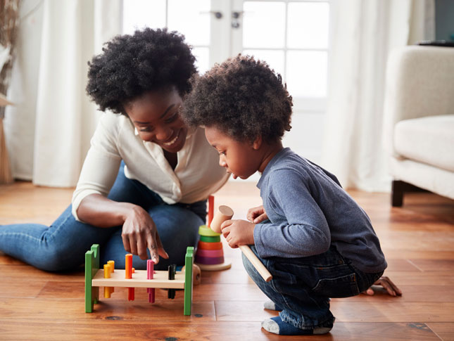 A mother smiles while sitting on the floor with her toddler son and playing with a toy.