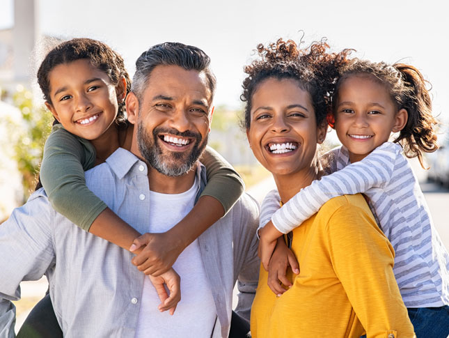 A father, mother and two daughters smile in an outdoor photo. Each parent has one of the daughters on their back.