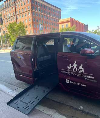 A wheel-chair accessible Toyota Sienna is parked at the curb with the ramp lowered. The adaptive van is burgundy and has Kennedy Krieger Institute's logo on the front passenger door. 