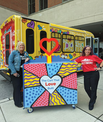 Two women smile as they stand on either side of a colorful cart that reads "The Cart That Love Built." There is a cutout of a red heart atop the cart.