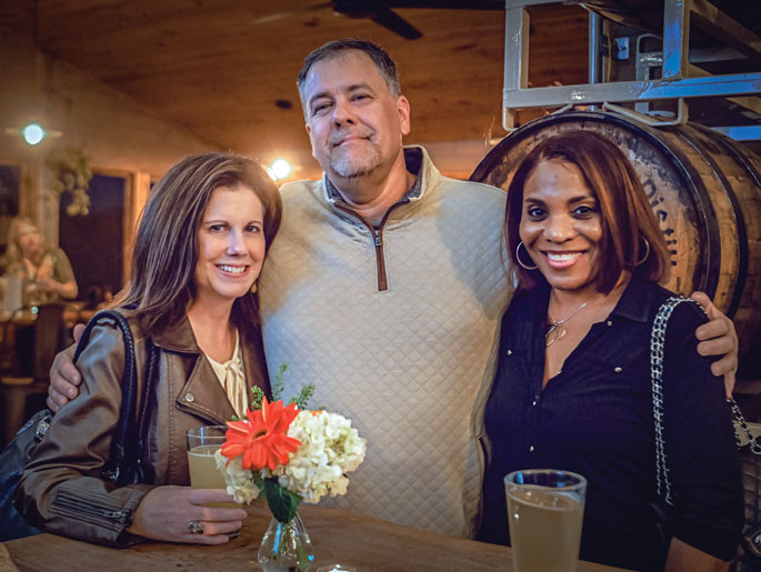 A man stands in between two women with his arms on their shoulder in a posed photo at Barrels & Bonfires. All three people are smiling.