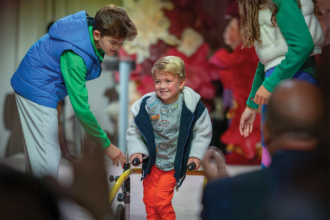 A young boy walks down the runway at Fall Fête 5 while using a mobility device. A boy and a woman are standing on either side of him to offer assistance.