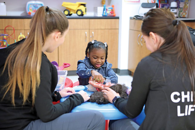 Two child life specialists sit at a table and play a game with a little girl. The little girl is facing the camera, while both child life specialists have their backs to the camera.