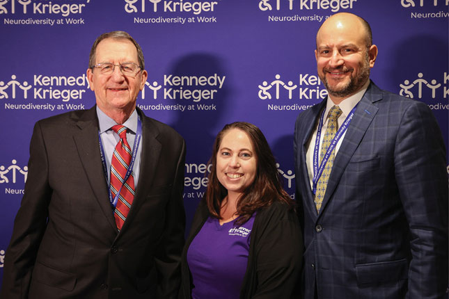 Two men and a woman stand in front of a purple and white step and repeat banner that features the Kennedy Krieger Neurodiversity at Work logo.