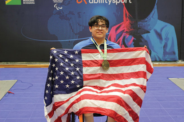 Noah Hassen sits in a mobility device while wearing a medal and holding the American flag. 