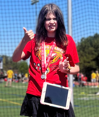 A girl wearing a augmentative and alternative communication device across her shirt stands up for a posed photo. She is participating in a Special Olympics event, and is also wearing a medal. 