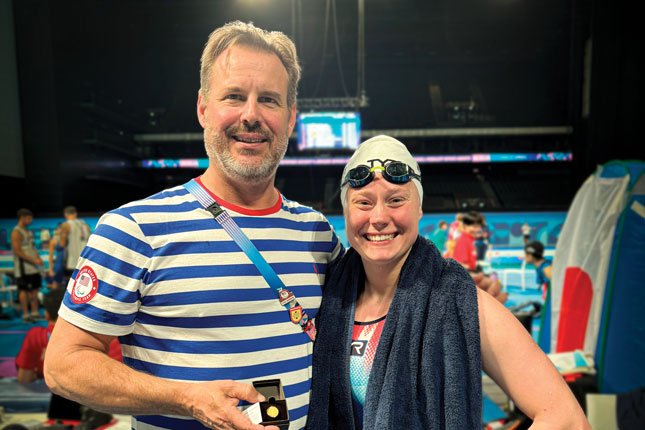 Taylor Winnett stands next to a man and smiles as the two stand poolside. She is wearing a swim cap and goggles.