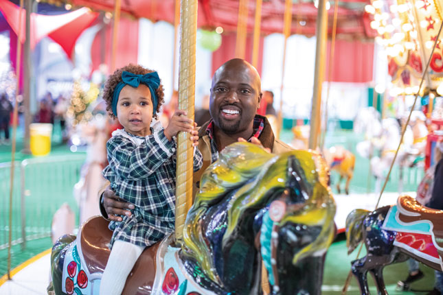 A little girl in a holiday dress rides a horse on a carousel at Kennedy Krieger's Festival of Trees. Her dad stands beside her, smiling.