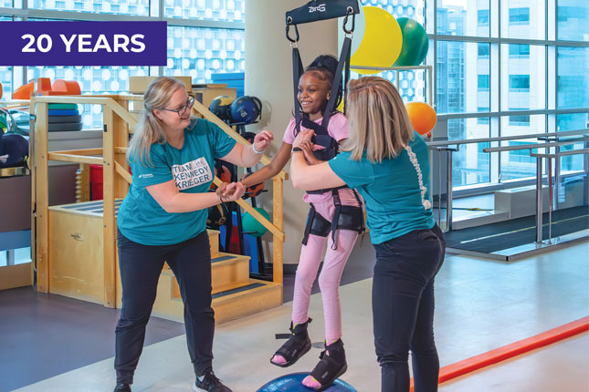A smiling young girl is suspended in a harness from a weight-supporting device. On her feet are black braces that support her feet and ankles. Helping her are two women, both wearing dark pants and teal T-shirts that say “Team Kennedy Krieger.” In the background are colorful therapy balls and other physical therapy equipment.