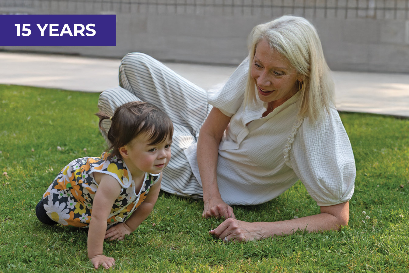 A very young girl crawls on the grass outside, while WIN co-president Cynthia Cavanaugh smiles beside her. Cavanaugh appears to be encouraging the girl to crawl.