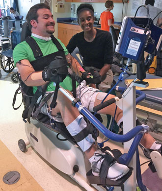 Ben Trockman smiles while using rehabilitative therapy equipment. He is strapped into a therapy chair, assisted by a therapist.