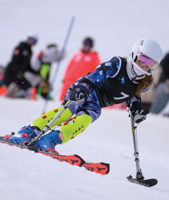 Victoria skis down a snowy hill wearing a helmet and ski suit, and holding ski poles, both of which are touching the snow and have forearm supports and wide bases.