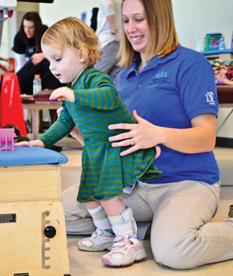 A little girl stands at a low therapy table with the help of a therapist behind her and braces on her lower legs.