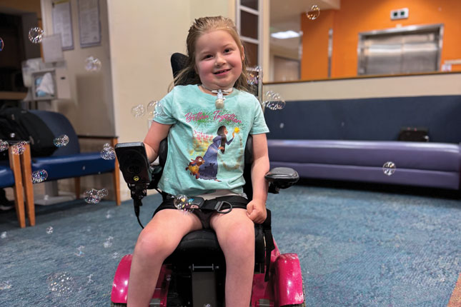 Lainey sits in the lobby of one of Kennedy Krieger's patient care buildings. She is smiling as bubbles float around her/